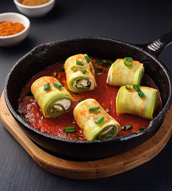 KI generiert: Das Bild zeigt eine Pfanne mit gefüllten, aufgerollten Zucchinischeiben in einer Tomatensauce, dekoriert mit Frühlingszwiebeln. Im Hintergrund sind Gewürzschalen zu sehen.