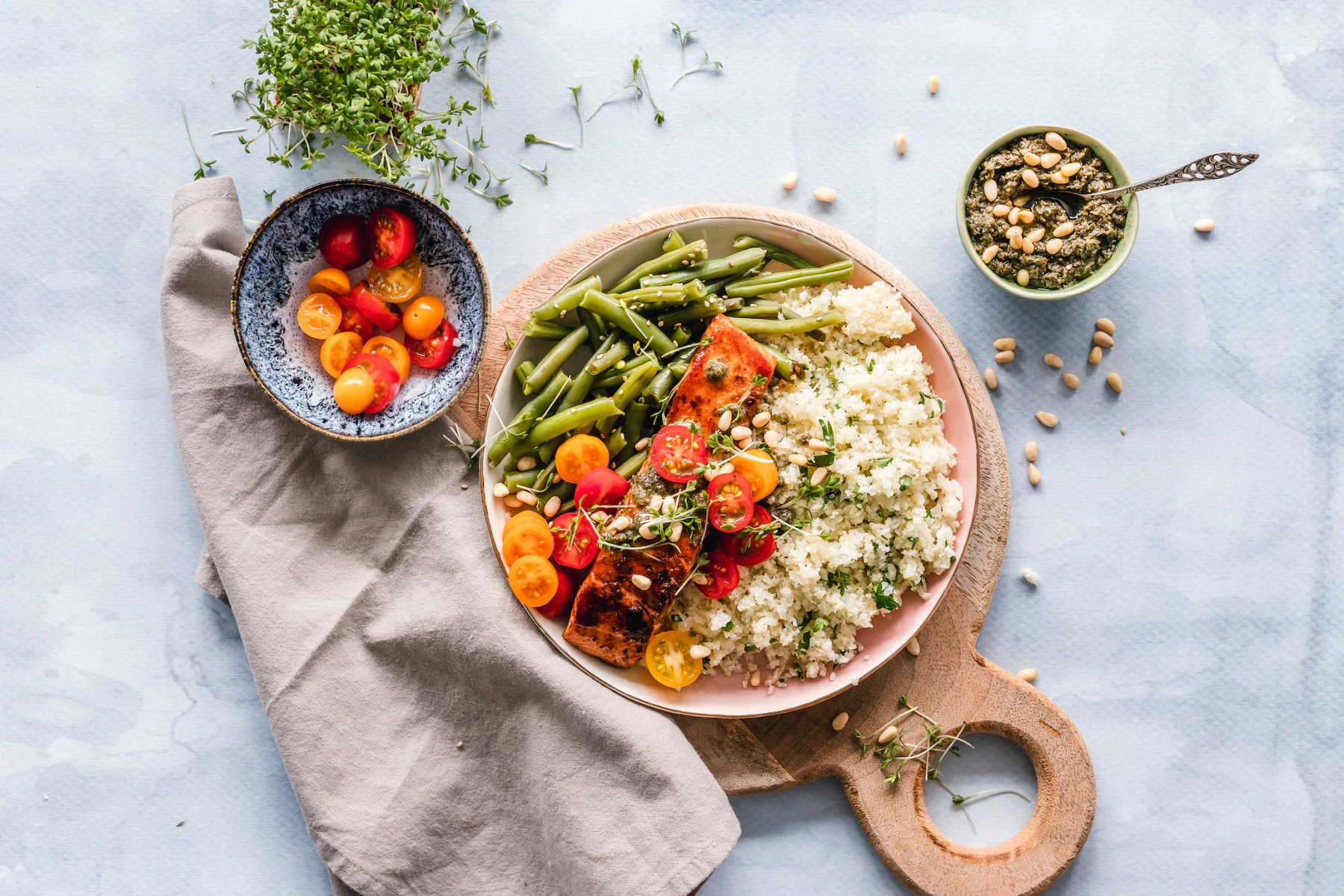 KI generiert: Das Bild zeigt ein reichhaltiges Gericht mit gebratenem Lachs, Quinoa, grünen Bohnen und frischen Tomaten, garniert mit Sprossen und Kernen. Daneben stehen zwei Schalen mit zusätzlichen Zutaten auf einem Holbrett, darunter Tomatenstücke und eine Gewürzmischung.