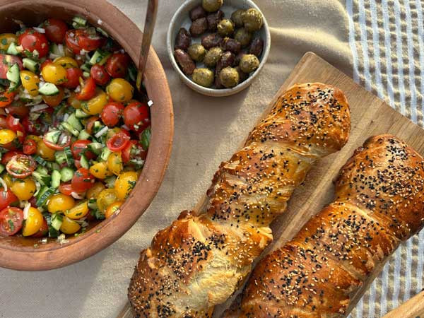 Auf einem Tisch steht ein Tomaten-Gurken-Salat mit 2 Baguettes und Oliven