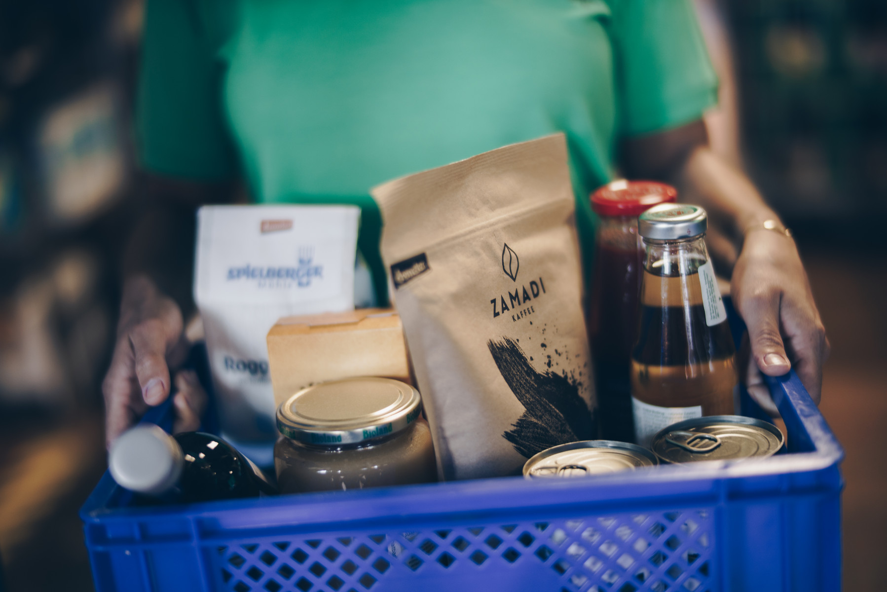 KI generiert: Das Bild zeigt eine Person, die einen blauen Korb mit verschiedenen Lebensmitteln trägt. Im Korb befinden sich verpackte Produkte wie Kaffee, Marmelade und Konserven.